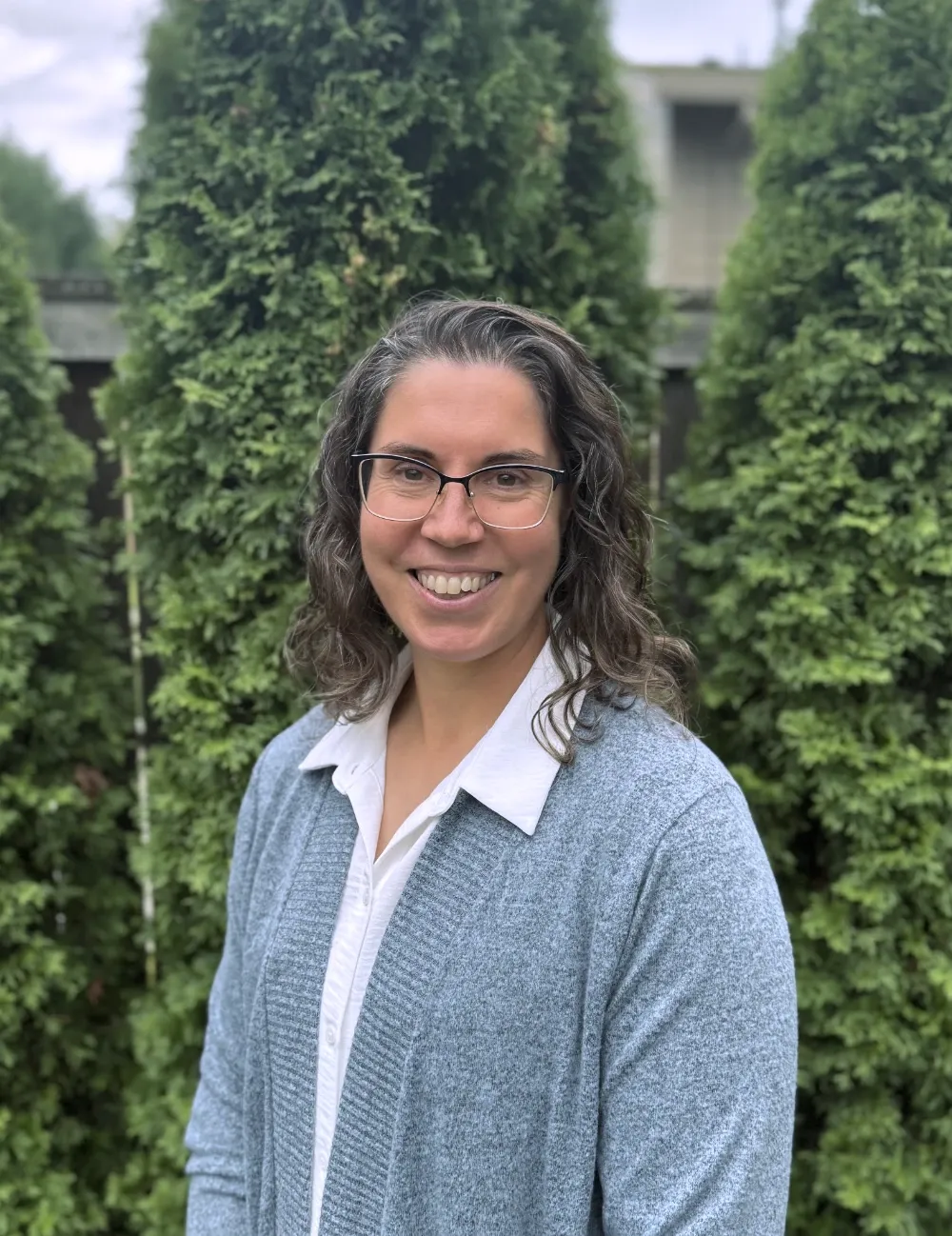 A picture of Abby, a woman in a blue cardigan standing in front of some greenery outdoors.