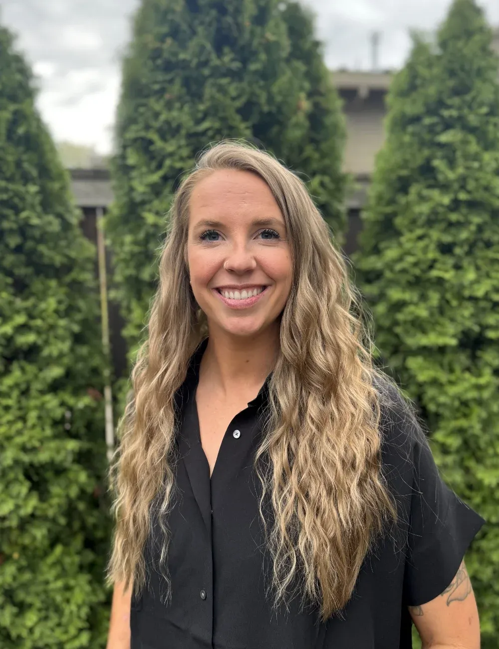 A picture of Andrea, a woman in a black shirt standing in front of some greenery outdoors.